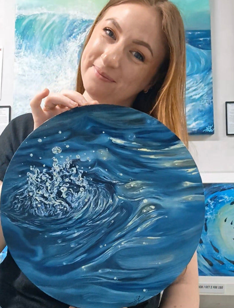 A woman holding a large round canvas painting of a blue ocean wave, with other ocean wave paintings visible on the wall behind her. Featuring HLSdesigners owner and artist, Hannah Singleton holding her recent oil painting, a drop in the ocean.