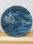 A round canvas painting depicting a swirling ocean wave in shades of deep blue, navy, and white, resting on a wooden surface against a light gray backdrop. Inspired by how the droplets fall from waves overhead, plunging into the surface of the ocean and firing up into the atmosphere, this is ‘a drop in the ocean’ an original oil painting, one of a kind, by Hannah singleton at HLSdesigner in Cornwall.