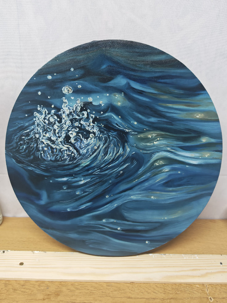 A round canvas painting depicting a swirling ocean wave in shades of deep blue, navy, and white, resting on a wooden surface against a light gray backdrop. Inspired by how the droplets fall from waves overhead, plunging into the surface of the ocean and firing up into the atmosphere, this is ‘a drop in the ocean’ an original oil painting, one of a kind, by Hannah singleton at HLSdesigner in Cornwall.