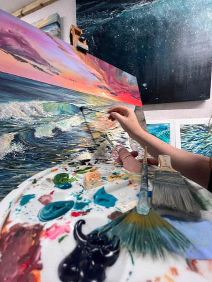close up painting using vibrant colours in acrylic, hand holding paintbrush and artists palette, focusing on original artwork of a beach scene, Hannah Singleton fine artist and illustrator from Cornwall is featured in the image as she creates her paintings inspired by the ocean 
