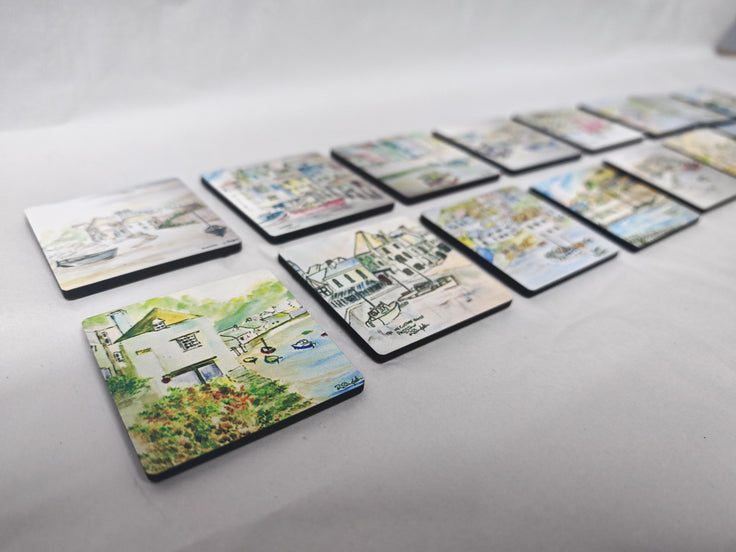 A set of square coasters with colourful coastal and harbour illustrations, arranged in a row on a light grey fabric background. Cornwall coastal scenes in watercolour by R Singleton reproduced exclusively by HLSdesigner into lovely collectable magnets, featuring the old watch house, Polperro, Sennen cove and more 