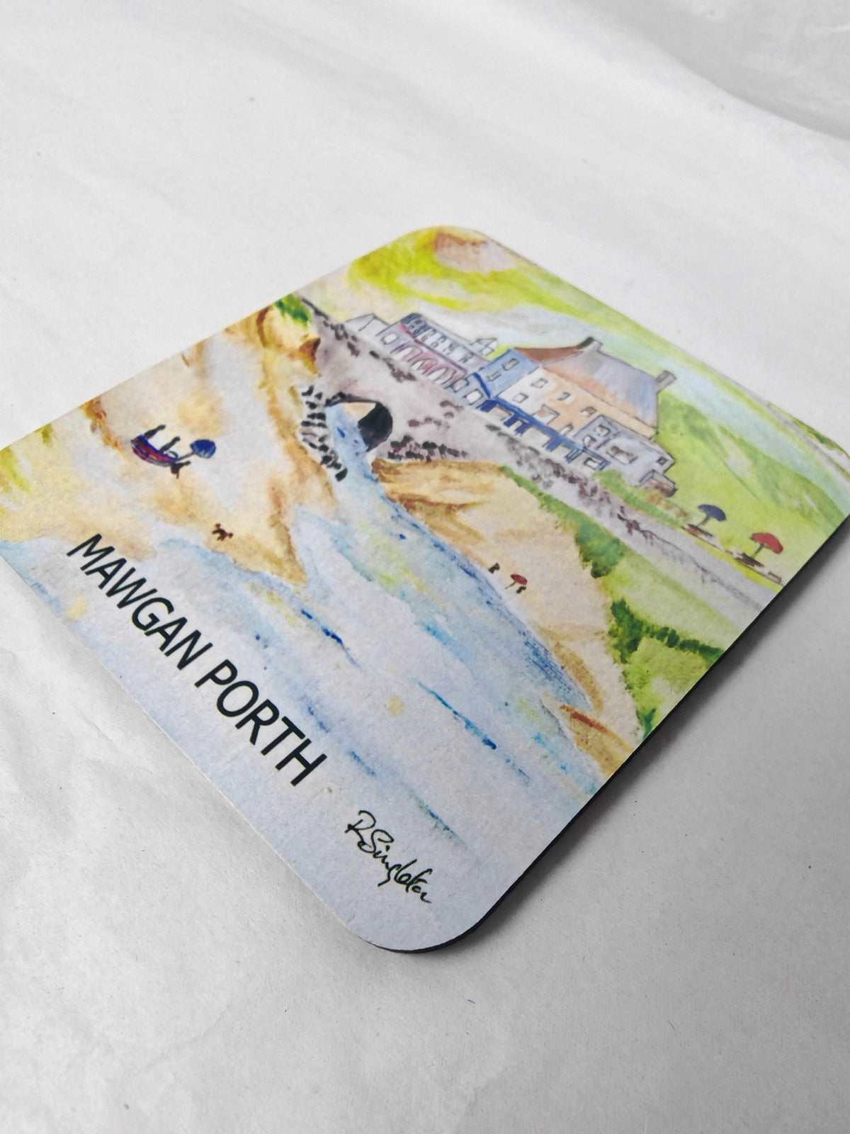 A square wooden coaster with rounded corners featuring Mawgan Porth beach near Newquay in Cornwall painted in watercolour by R Singleton focusing on the Merrymoor inn pub and the bridge where the stream flows under towards the ocean with beach goers on the sand. Printed exclusively at HLSdesigner