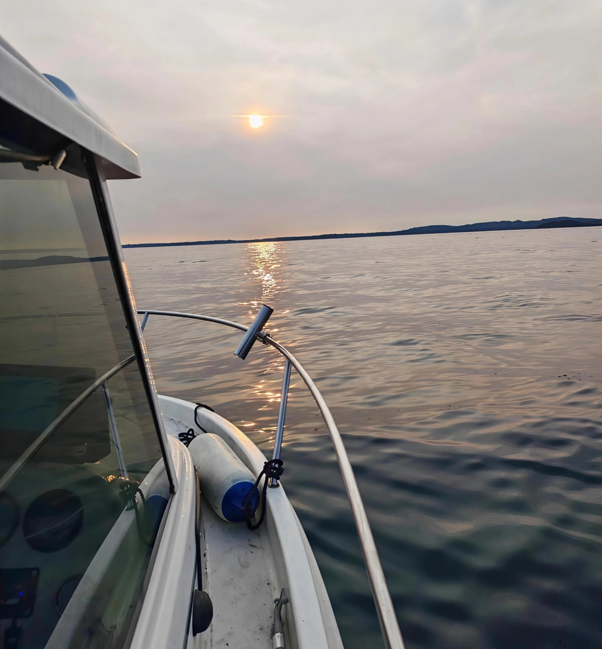 A close-up view from the bow of a white boat on open water during sunset, with calm ripples, distant land, and the sun reflecting on the water. Taken by HLSdesigner at Fowey harbour, Cornwall.