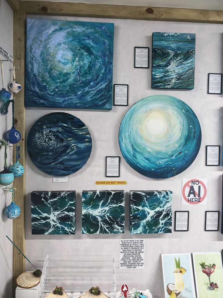 A gallery-style wall display featuring multiple ocean-wave themed artworks, including several square and circular paintings in swirling blue, teal, and white tones, with small framed labels and a 'No AI Art' sign nearby. All of my work is painted and designed by me, Hannah Singleton at HLSdesigner in Cornwall. Featuring ‘a drop in the ocean’ original oil painting inspired by close up viewpoints of the ocean.