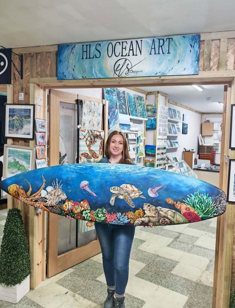 Artist, Hannah Singleton stands in her shop doorway holding a large, colourful surfboard painted with an ocean scene featuring a sea turtle, jellyfish, and coral reef. A wooden sign above the doorway reads 'HLS OCEAN ART by HLSdesigner'. This is the Beneath the break original surfboard. Newquay, Cornwall, ocean inspired artwork, original art, prints and home décor inspired by coastal viewpoints of the ocean and Cornwall scenes. 