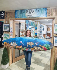 Artist, Hannah Singleton stands in her shop doorway holding a large, colourful surfboard painted with an ocean scene featuring a sea turtle, jellyfish, and coral reef. A wooden sign above the doorway reads 'HLS OCEAN ART by HLSdesigner'. This is the Beneath the break original surfboard. Newquay, Cornwall, ocean inspired artwork, original art, prints and home décor inspired by coastal viewpoints of the ocean and Cornwall scenes. 