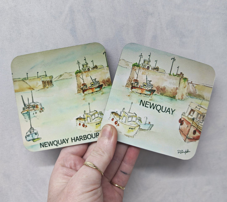 Two square coasters with rounded corners featuring a watercolor illustration of Newquay Harbour and boats, held in a hand against a textured light gray background. This is the Newquay harbour watercolour print by R Singleton reproduced onto coasters showing the whole scene across two collectable coaster souvenir gifts. By R Singleton and printed by HLSdesigner hlsoceanart cornwall.
