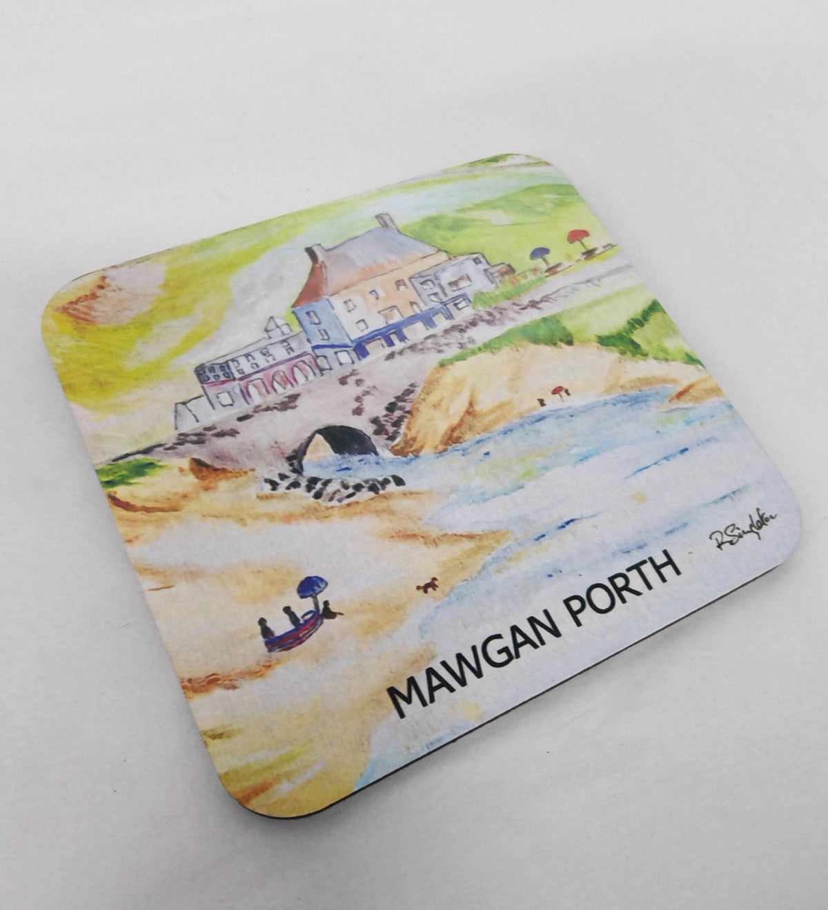 A square printed coaster featuring a colorful watercolor illustration of Mawgan Porth beach and building, with the text 'MAWGAN PORTH' printed at the bottom, photographed on a white fabric surface. Painted in watercolour by R Singleton and exclusive to HLSdesigner