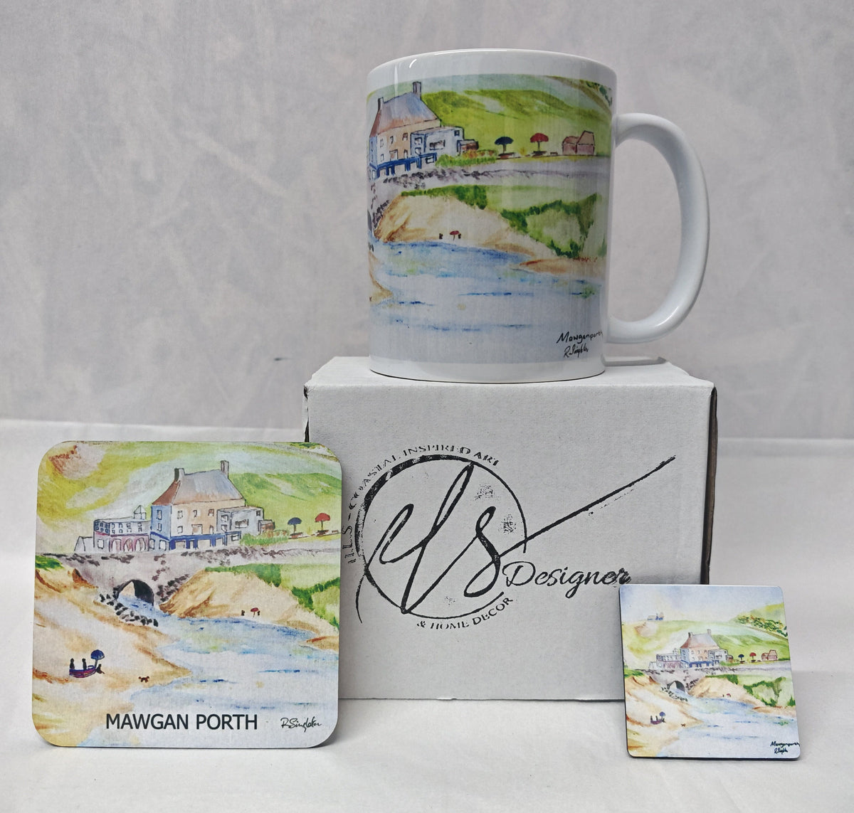 A white ceramic mug with a watercolour coastal scene stands on top of a white box, with matching printed coasters displayed in front. The box and coasters show the brand name 'HLS Designer' and the location 'Mawgan Porth'. Painted in watercolour by R Singleton and reproduced by his daughter Hannah in tribute. In the collection is a mug, a coaster and a magnet as well as available fine art prints reproduced from his original Cornwall paintings