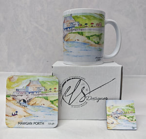 A white ceramic mug with a watercolour coastal scene stands on top of a white box, with matching printed coasters displayed in front. The box and coasters show the brand name 'HLS Designer' and the location 'Mawgan Porth'. Painted in watercolour by R Singleton and reproduced by his daughter Hannah in tribute. In the collection is a mug, a coaster and a magnet as well as available fine art prints reproduced from his original Cornwall paintings