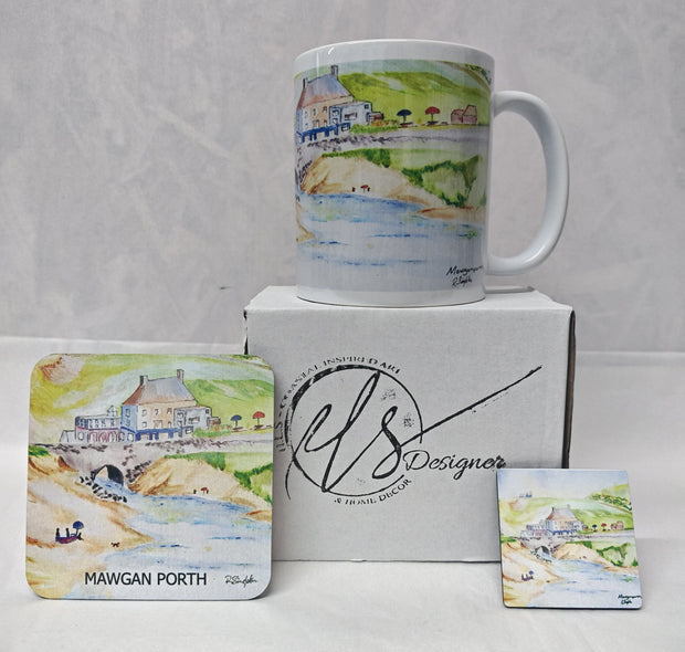 A white ceramic mug with a watercolour coastal scene stands on top of a white box, with matching printed coasters displayed in front. The box and coasters show the brand name 'HLS Designer' and the location 'Mawgan Porth'. Painted in watercolour by R Singleton and reproduced by his daughter Hannah in tribute. In the collection is a mug, a coaster and a magnet as well as available fine art prints reproduced from his original Cornwall paintings