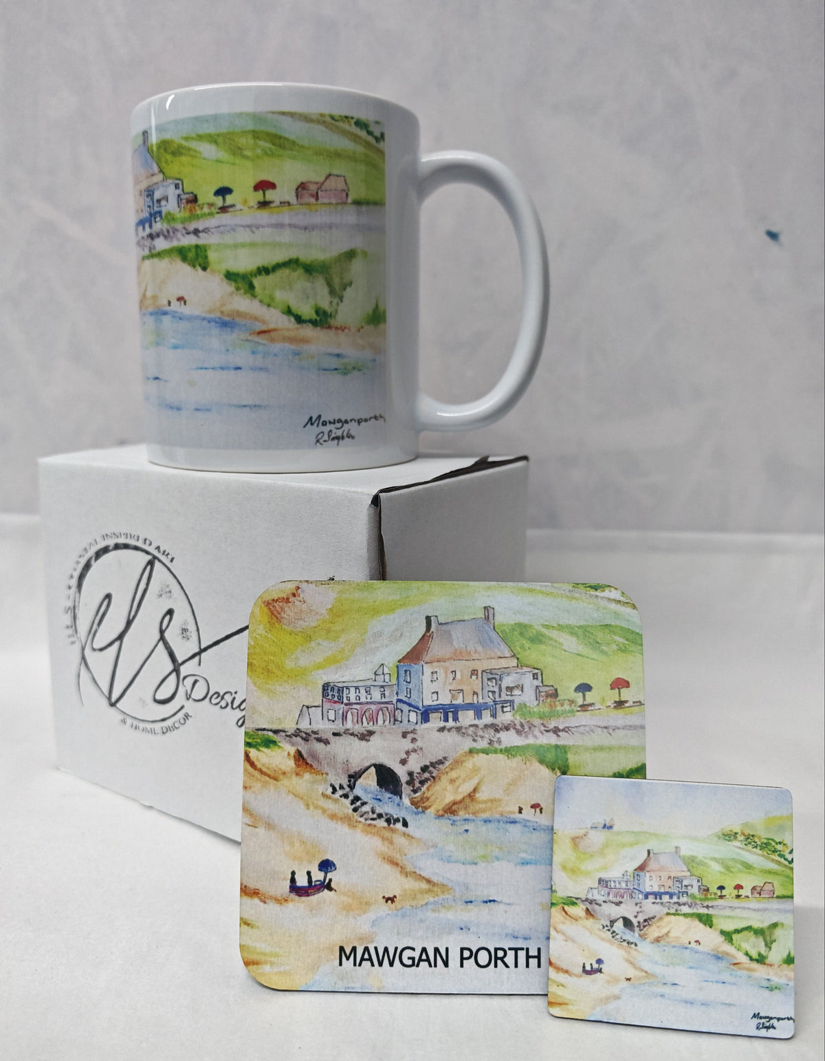 A white ceramic mug and matching coaster and magnet featuring a watercolour illustration of Mawgan Porth beach, displayed on a white box against a light Gray background. HLSdesigner has reproduced these prints on to accessories in tribute to the late R Singleton coastal Cornwall scene artist 