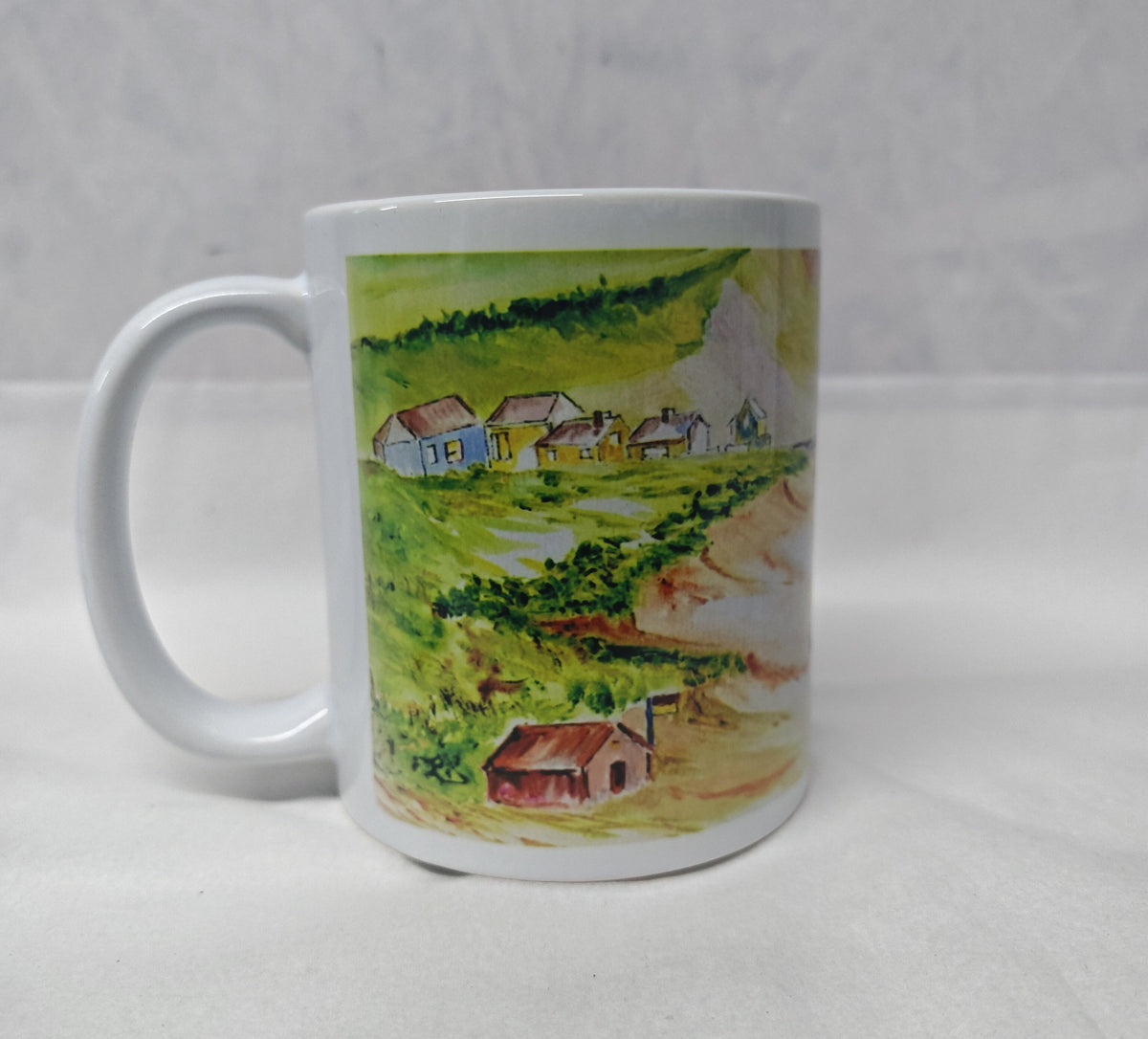 A white ceramic mug featuring a printed landscape illustration of a small village with green hills, trees, and houses, set against a light Gray textured background. This is the side of the Mawgan Porth beach painting in watercolour by R Singleton - reproduced onto a mug and other accessories exclusive to HLSdesigner in Cornwall. Coastal seascape beach paintings 