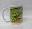 A white ceramic mug featuring a printed landscape illustration of a small village with green hills, trees, and houses, set against a light Gray textured background. This is the side of the Mawgan Porth beach painting in watercolour by R Singleton - reproduced onto a mug and other accessories exclusive to HLSdesigner in Cornwall. Coastal seascape beach paintings 