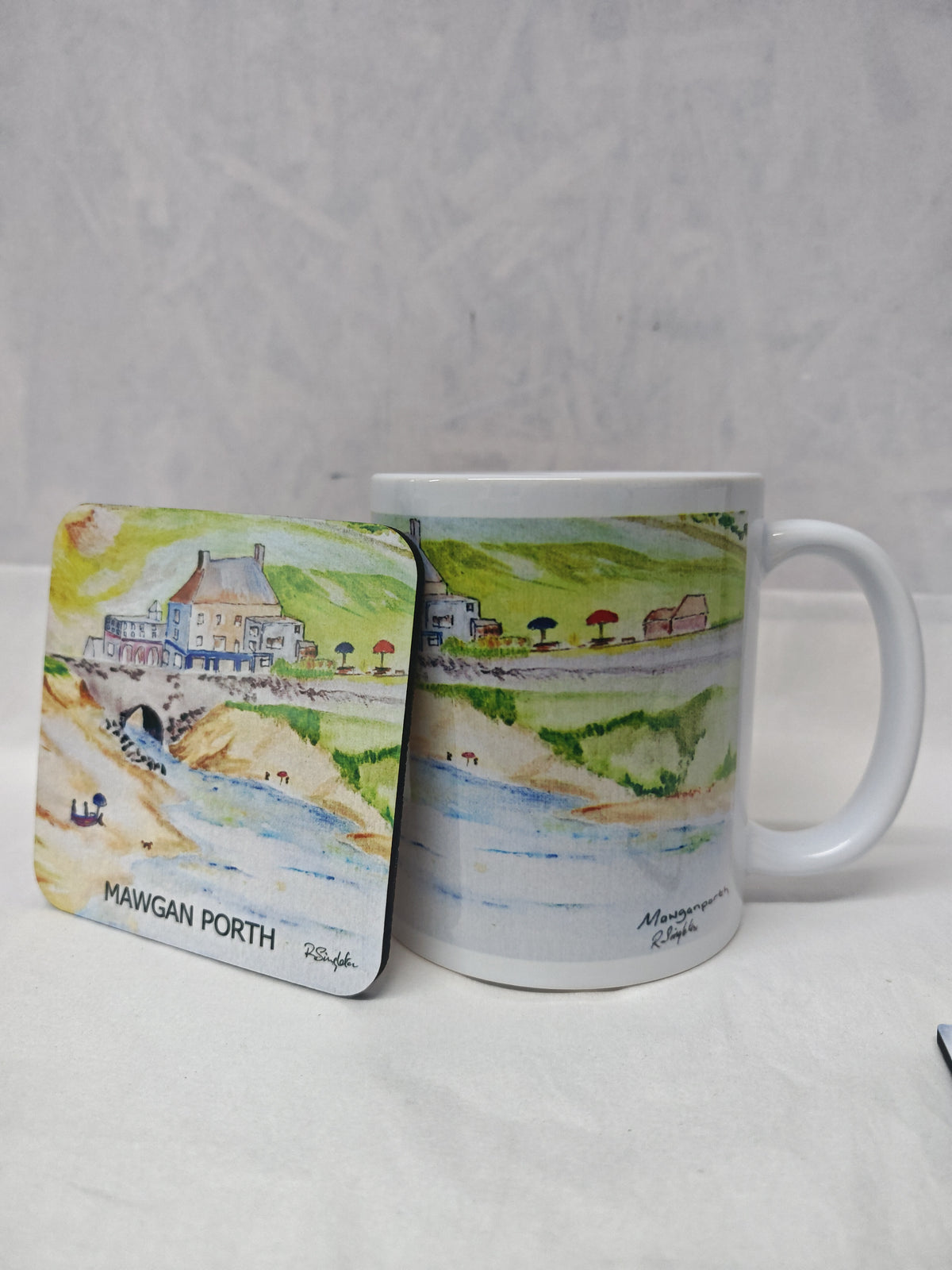 A white ceramic mug and matching square coaster printed with a watercolor landscape of Mawgan Porth, shown on a light gray textured background.