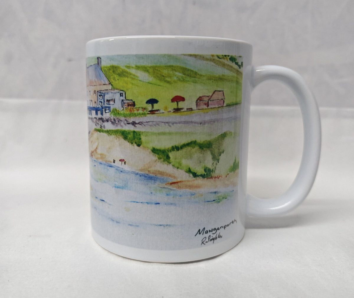 A white ceramic coffee mug featuring a watercolour landscape illustration of a coastal village with green hills, blue water, and small buildings. The mug is photographed against a neutral, light grey textured background. This is a watercolour painting by R Singleton of Mawgan Porth beach with the Merrymoor inn beside it, featuring washes of colour and a modern depiction of sketch work , reproduced y HLSdesigner in Cornwall