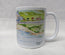 A white ceramic coffee mug featuring a watercolour landscape illustration of a coastal village with green hills, blue water, and small buildings. The mug is photographed against a neutral, light grey textured background. This is a watercolour painting by R Singleton of Mawgan Porth beach with the Merrymoor inn beside it, featuring washes of colour and a modern depiction of sketch work , reproduced y HLSdesigner in Cornwall