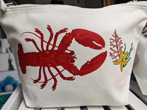 red lobster illustration by hlsdesigner printed and pressed in house onto beautiful cosmetic accessory bags, this one features the medium bag close up with a yellow starfish, seaweed and coral bundle accent on a white shelf and is complete with a silver metal zip.