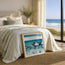 A framed art print of ocean waves leaning on a bed in a beautifully styled bedroom with a large window overlooking the beach and ocean. This is Dusk Tide - a representation of a wave crashing into the shore at dusk with a foggy sky and clear white seafoam. Painted and designed by Hannah Singleton at HLSdesigner, coastal fine art based in Cornwall inspired by the Cornish coastline