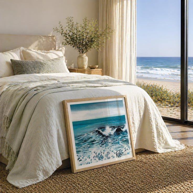 A framed art print of ocean waves leaning on a bed in a beautifully styled bedroom with a large window overlooking the beach and ocean. This is Dusk Tide - a representation of a wave crashing into the shore at dusk with a foggy sky and clear white seafoam. Painted and designed by Hannah Singleton at HLSdesigner, coastal fine art based in Cornwall inspired by the Cornish coastline