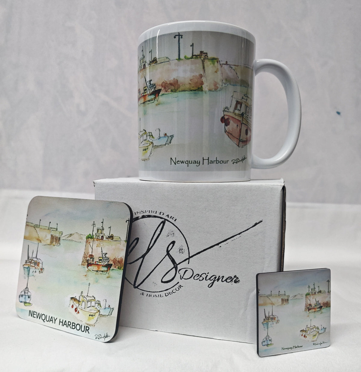 A white ceramic mug and two matching square coasters printed with a watercolour Newquay Harbour scene, displayed with a branded white gift box on a white fabric surface. watercolour painting of Newquay harbour on a ceramic mug as well as a wooden coaster and magnet with hand printed mug box - painted by R Singleton Cornwall coastal scenes and reproduced by HLSdesigner