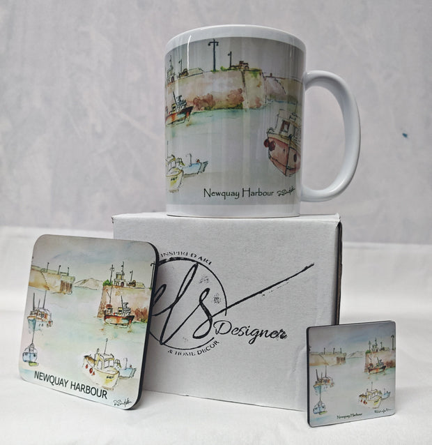 A white ceramic mug and two matching square coasters printed with a watercolour Newquay Harbour scene, displayed with a branded white gift box on a white fabric surface. watercolour painting of Newquay harbour on a ceramic mug as well as a wooden coaster and magnet with hand printed mug box - painted by R Singleton Cornwall coastal scenes and reproduced by HLSdesigner