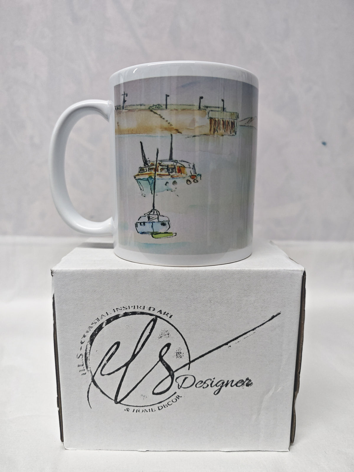 A white ceramic mug with a watercolour illustration of boats on water, displayed on top of its white branded box. HLSdesigner. painting by R Singleton as tribute printed onto mugs and accessories, Newquay Harbour Cornwall 