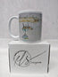 A white ceramic mug with a watercolour illustration of boats on water, displayed on top of its white branded box. HLSdesigner. painting by R Singleton as tribute printed onto mugs and accessories, Newquay Harbour Cornwall 