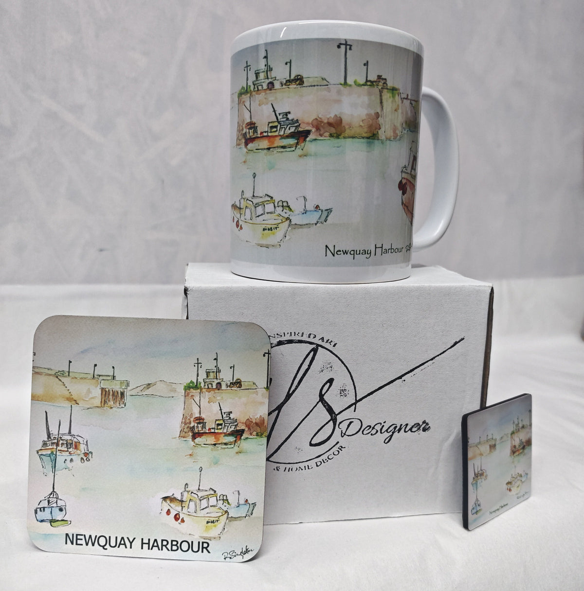 A Newquay Harbour themed gift set featuring a white ceramic mug, a square coaster, a small square wooden magnet to match and a branded HLSdesigner white box. The items are decorated with a watercolour painting of fishing boats on a harbour and the text 'Newquay Harbour' and 'Newquay Bay'.