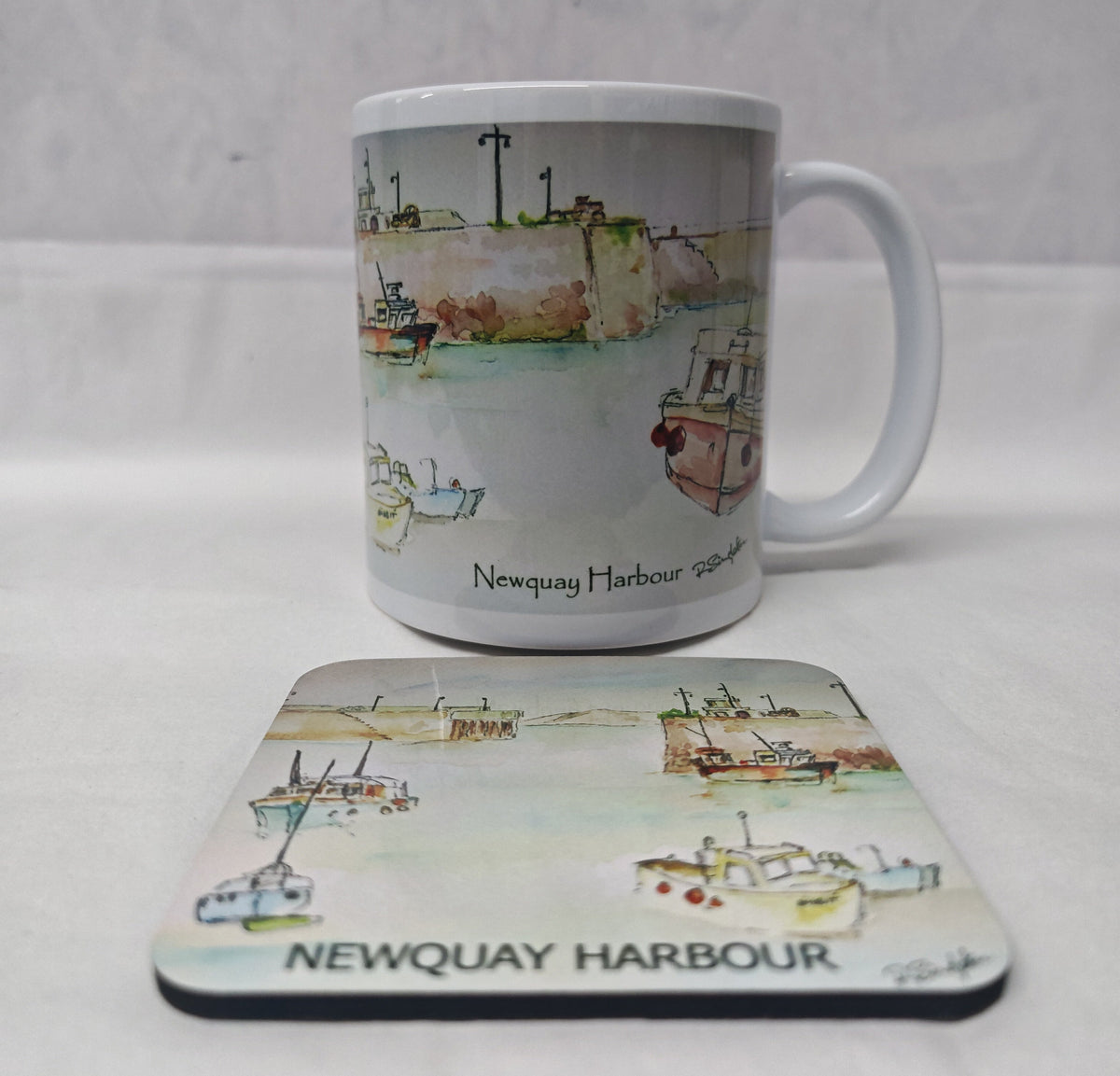A white ceramic mug and matching rectangular coaster featuring a watercolour illustration of Newquay Harbour with boats and the text 'Newquay Harbour' and 'NEWQUAY HARBOUR'. HLSdesigner
