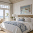 A beautifully styled beach-themed bedroom featuring a large bed with white linens, a blue and white leaf-patterned throw blanket, and a woven rattan headboard. A framed coastal art print of Newquay harbour hangs on the wall above the bed, and a glass door on the left offers a view of the ocean and palm tree. The painting features studies of the fishing boats that are local to the harbour, coming in after days of fishing and working aboard. A contemporary style shows the modern day while keeping it historic
