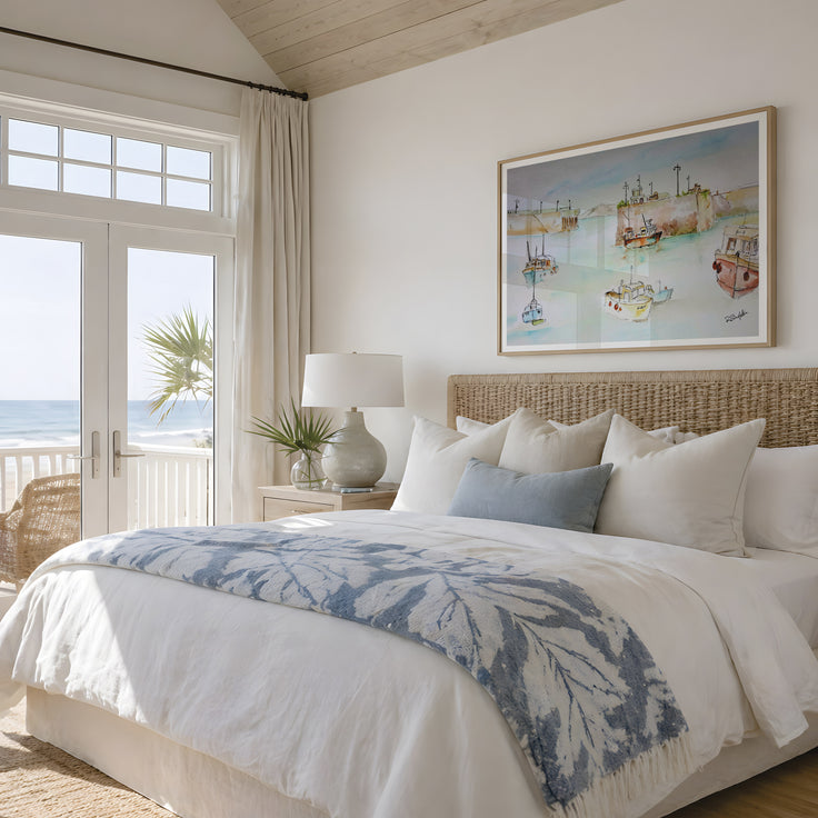 A beautifully styled beach-themed bedroom featuring a large bed with white linens, a blue and white leaf-patterned throw blanket, and a woven rattan headboard. A framed coastal art print of Newquay harbour hangs on the wall above the bed, and a glass door on the left offers a view of the ocean and palm tree. The painting features studies of the fishing boats that are local to the harbour, coming in after days of fishing and working aboard. A contemporary style shows the modern day while keeping it historic