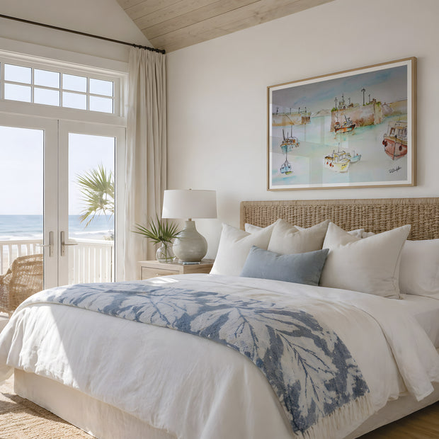 A beautifully styled beach-themed bedroom featuring a large bed with white linens, a blue and white leaf-patterned throw blanket, and a woven rattan headboard. A framed coastal art print of Newquay harbour hangs on the wall above the bed, and a glass door on the left offers a view of the ocean and palm tree. The painting features studies of the fishing boats that are local to the harbour, coming in after days of fishing and working aboard. A contemporary style shows the modern day while keeping it historic