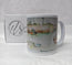 A white ceramic mug featuring a watercolour illustration of Newquay Harbour with boats and a quay wall, shown next to its white branded gift box on a white fabric background. painted in watercolour by R Singleton Cornwall artist and reproduced by HLSdesigner