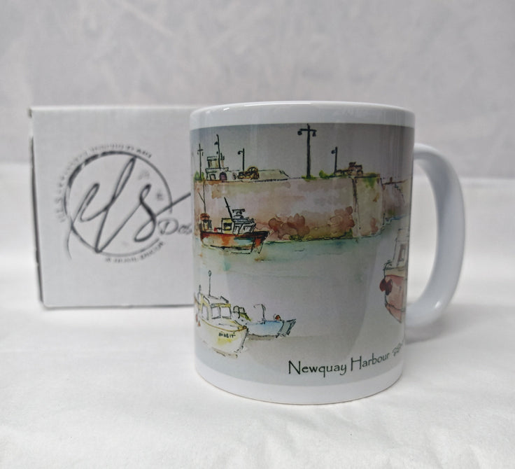 A white ceramic mug featuring a watercolour illustration of Newquay Harbour with boats and a quay wall, shown next to its white branded gift box on a white fabric background. painted in watercolour by R Singleton Cornwall artist and reproduced by HLSdesigner