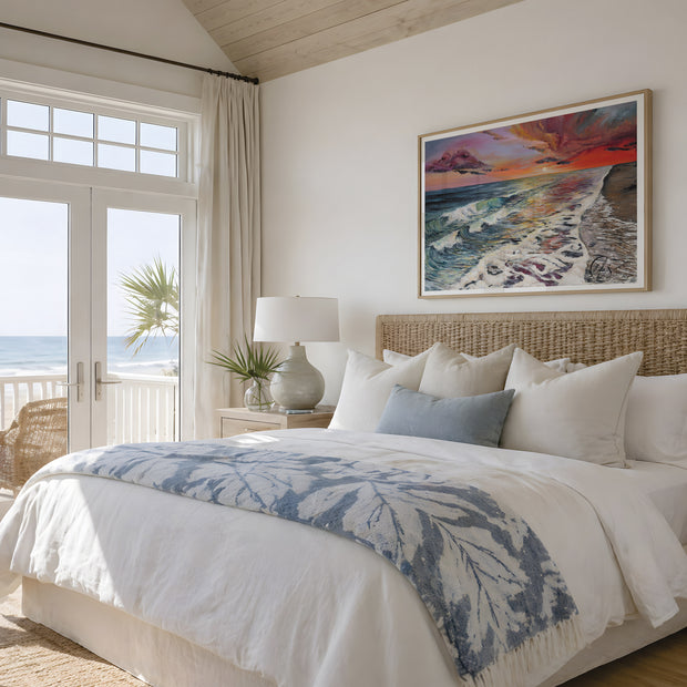 A beautifully styled beach-themed bedroom featuring a large bed with white linens, a blue and white patterned throw blanket, and a wicker headboard. A colorful framed painting of an ocean sunset hangs on the wall above the bed, and a glass door on the left offers a view of the ocean. The ocean positioned to the left crashes into the shoreline leaving seafoam and coloured reflections of the piercing red sunset from above. Originally painted by Hannah Singleton at HLSdesigner, ocean artist based in Cornwall. 