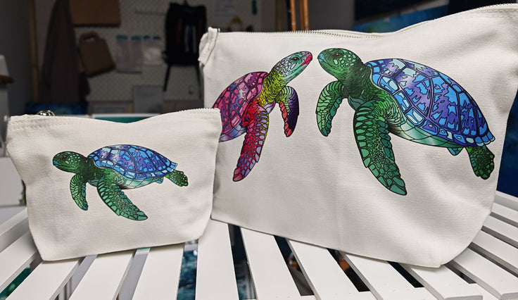 printed and designed in house, these accessory bags are sitting next to each other to compare in size, showing the turtle design collection. sitting on a white shelf with ocean art in the background