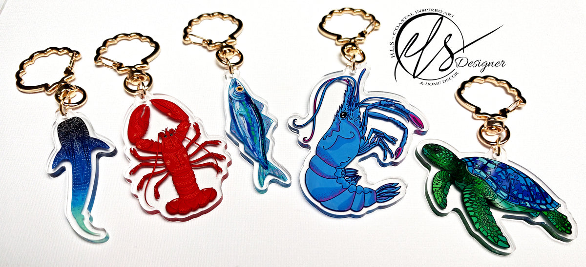 Set of colourful keychains shaped like a whale shark, lobster, fish, and turtle on a white background. Featuring drawn designs by HLSdesigner in Cornwall, Green sea turtle, blue king shrimp with pink detailing and spotted shading, blue watercolour inspired sardine with orange eye, red and orange spotted king lobster and a constellation inspired whale shark illustration with spots, linear work and stars. All printed into acrylic and finished with a gold metal clasp in the shape of a seashell.
