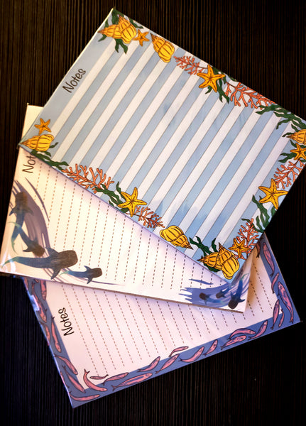 handmade and designed by HLSdesigner, three 50 page tear off  notepads ideal for all notes and list making. the top one incorporates seashells, coral, seaweed and starfish on a striped lined paper background in blue and white. the middle notepad has beautiful whale sharks swimming around the page with a dotted inner for note taking and brush watercolour waves around it on a white background, the third and bottom notepad is the fish shoal pad of fish swimming around the page in pinks and purples