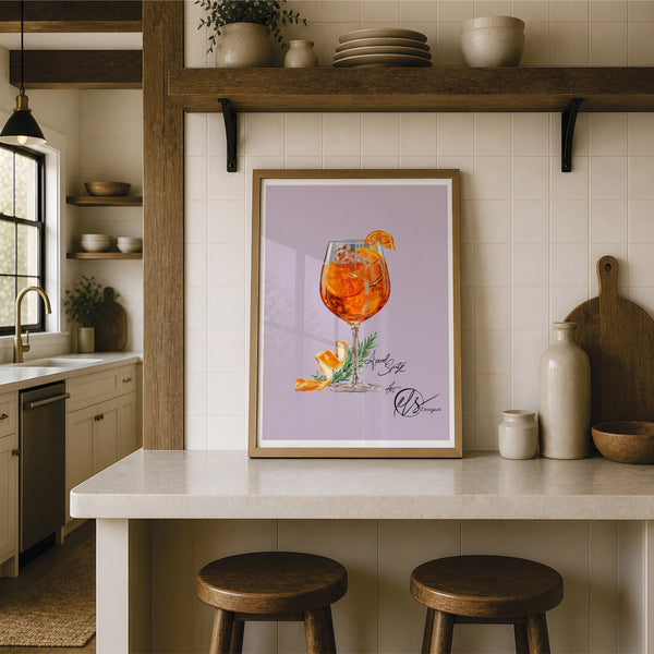 A framed art print featuring an illustration of an orange aperol spritz cocktail in a glass with an orange slice garnish, displayed on a kitchen counter in a rustic kitchen setting. A stylized illustration of an orange cocktail in a stemmed glass with orange slices, ice, and a citrus peel garnish, set on a light lavender background with repeated watermark text and the signature 'HLS Designer' at the bottom right. 