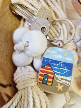 Keychain with a colourful design of a beach house and starfish in front of the ocean on a sunny day, surrounded by seashells and rope.
