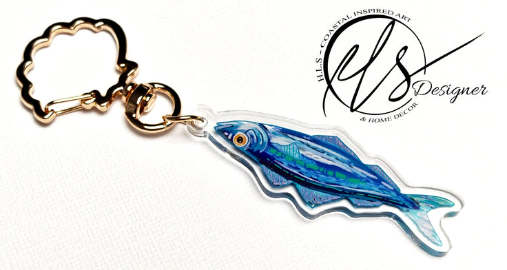 watercolour inspired sardine illustration with blues and greens throughout and a bold orange fish eye. Finished in clear acrylic and shaped to the fish with a metal seashell shaped keychain clasp for ocean decoration, image taken on a white background with the HLSdesigner logo in the corner, coastal inspired art and home décor 