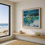 A framed square ocean painting hangs on a beige wall in a modern room with a window showing a beach view, a wooden shelf, and decorative seashells. The print on the wall is a fine art print by HLSdesigner of a crashing wave with green and yellow hues shining through the crest as the wave barrels over into the shore. Reflecting the sunlight above it. The painting has the canvas texture throughout the brush strokes and this is also represented within the print. Designed by HLSdesigner, Hannah Singleton