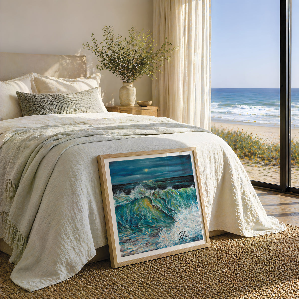 A framed painting of a dramatic ocean wave with blue and green tones leans against a bed in a bright, modern bedroom. The room has cream bedding, a vase with branches on a nightstand, and a large window overlooking a sandy beach and ocean. The fine art print is represented not to scale. By Cornwall based fine artist Hannah Singleton at HLSdesigner
