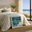 A framed painting of a dramatic ocean wave with blue and green tones leans against a bed in a bright, modern bedroom. The room has cream bedding, a vase with branches on a nightstand, and a large window overlooking a sandy beach and ocean. The fine art print is represented not to scale. By Cornwall based fine artist Hannah Singleton at HLSdesigner