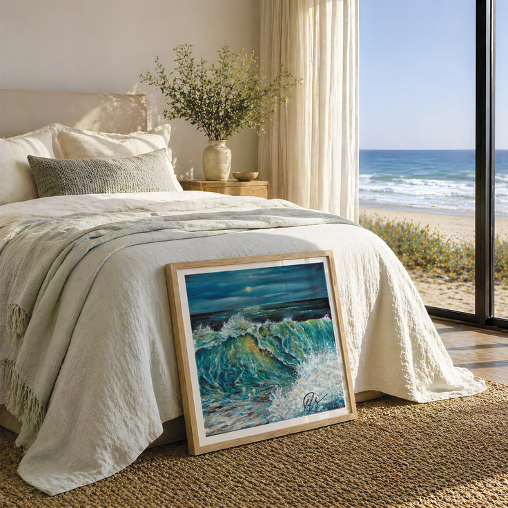 A framed painting of a dramatic ocean wave with blue and green tones leans against a bed in a bright, modern bedroom. The room has cream bedding, a vase with branches on a nightstand, and a large window overlooking a sandy beach and ocean. The fine art print is represented not to scale. By Cornwall based fine artist Hannah Singleton at HLSdesigner
