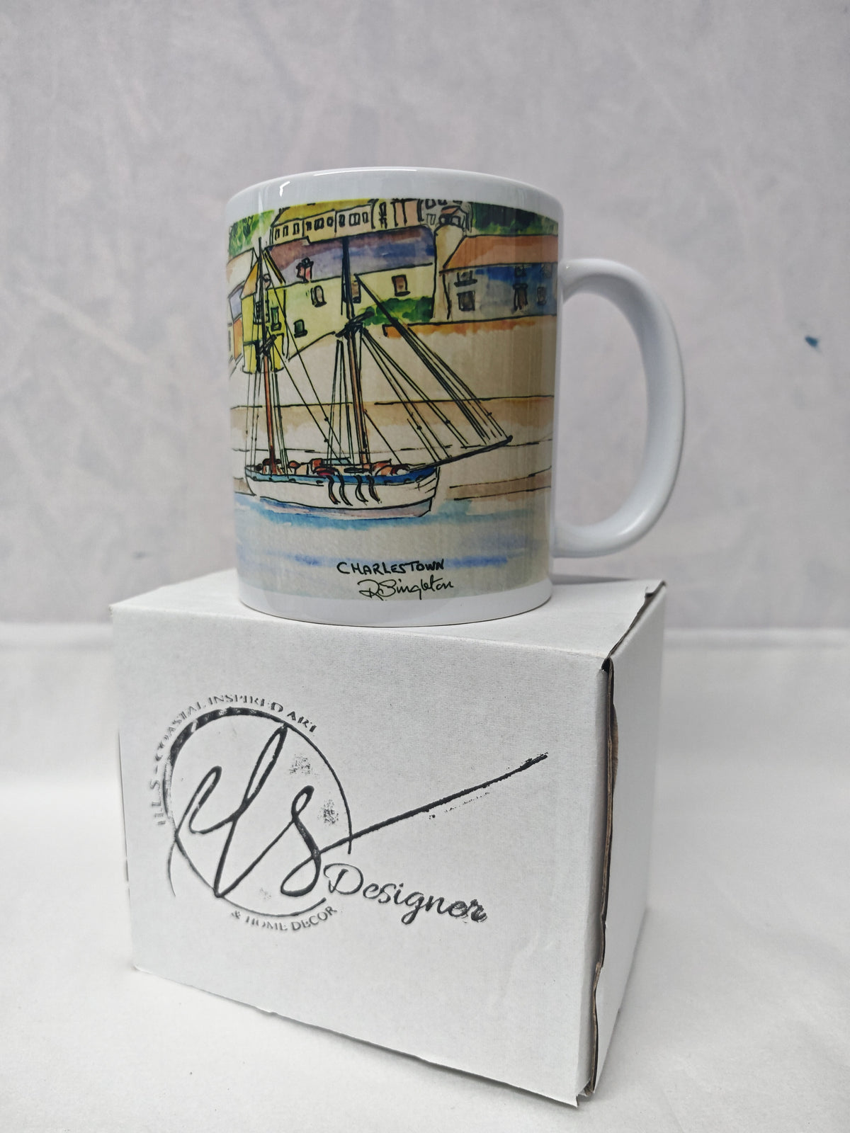 A white ceramic mug featuring a watercolour painting of Charlestown harbour in cornwall by artist R Singleton - reproduced in tribute by his daughter exclusively at HLSdesigner onto accessories and collectable prints. Focusing on the tall ships moored against the quay wall and the fishing village in the background, positioned onto a HLSdesigner branded mug box on a white background