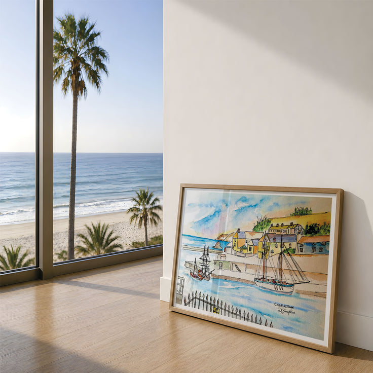 A framed watercolour painting of a coastal harbour scene with boats, palm trees, and a beach is leaning against a white wall on a light wood floor, with a large window showing the ocean and sky behind it. This is Charlestown - an old historic fishing port in Cornwall famous for its regatta and tall ships. This painting was originally created by Robert Singleton and has been lovingly reproduced by HLSdesigner. 