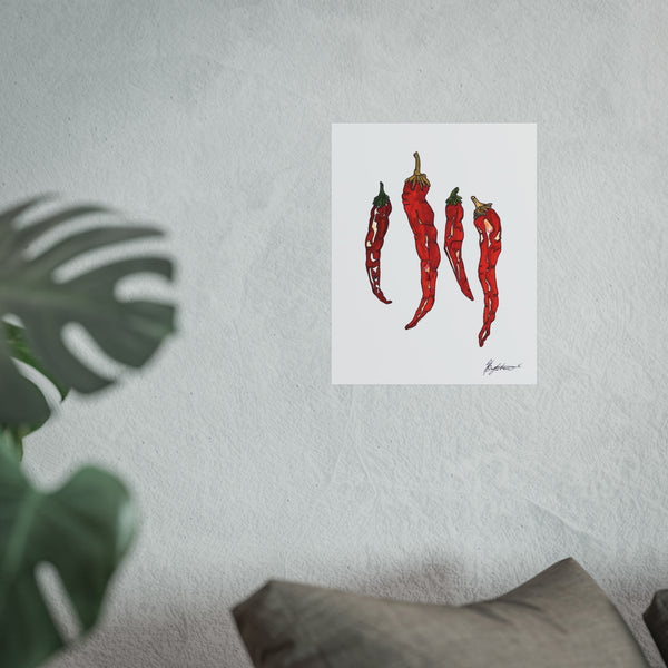 Art print of red chili peppers on a white background, leaning against a textured wall with a plant and pillow in the foreground.