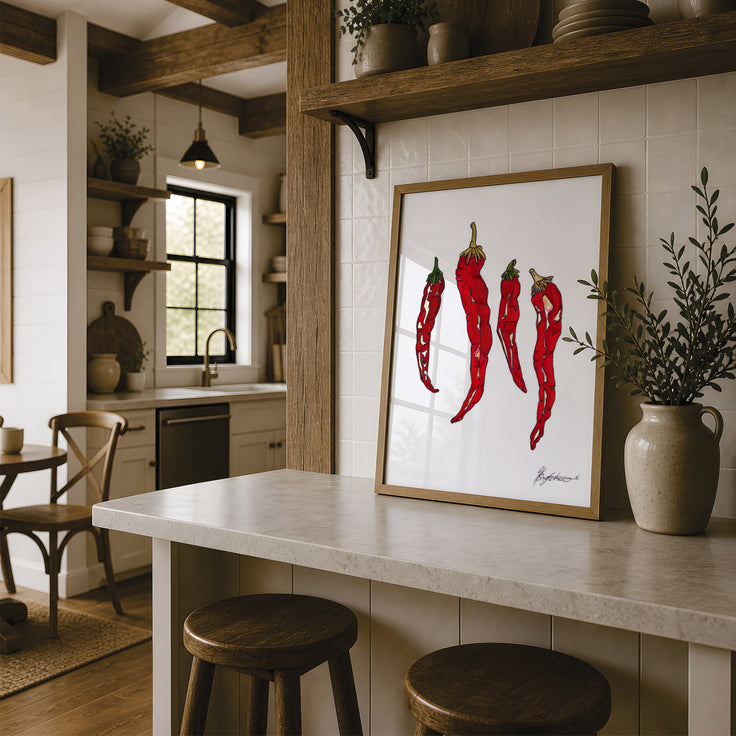 A framed art print featuring three red chili peppers on a white background, styled on a kitchen island in a rustic kitchen setting with wooden stools, a stone countertop, and a ceramic vase. Fine art print by Hannah Singleton at HLSdesigner using watercolour and mixed media